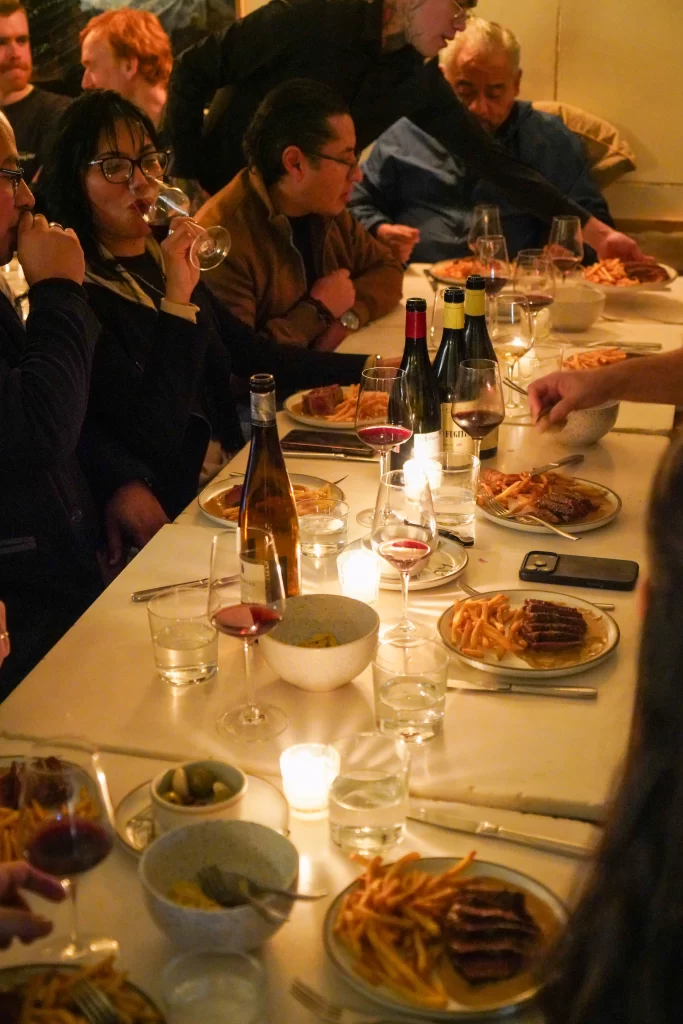 La mesa de la cena de bienvenida, con el plato principal y las copas. Algunas botellas de vino con invitados sonriendo y tomando.